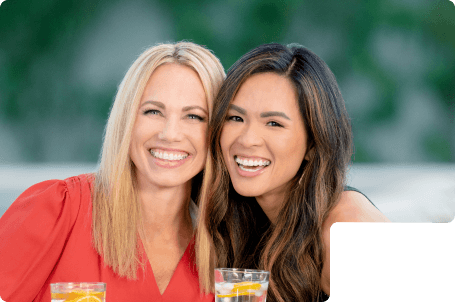 2 adult women smiling with straight white teeth after finishing Clear Braces treatment.