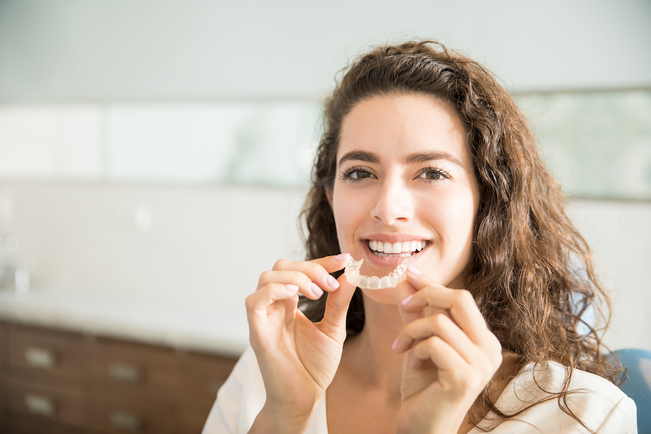 A smiling lady holding a clear aligner tray in front of her mouth.