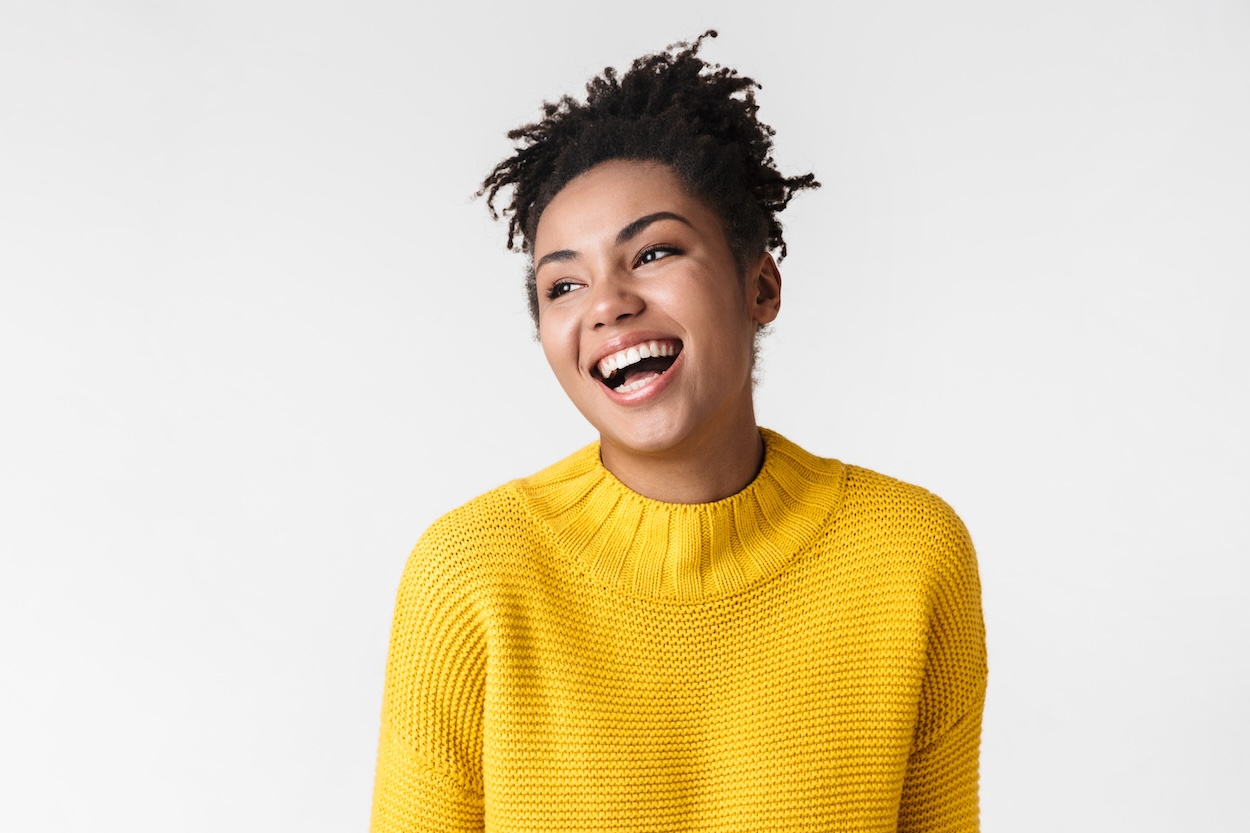 Happy smiling young woman wearing a yellow sweater with a grey background.