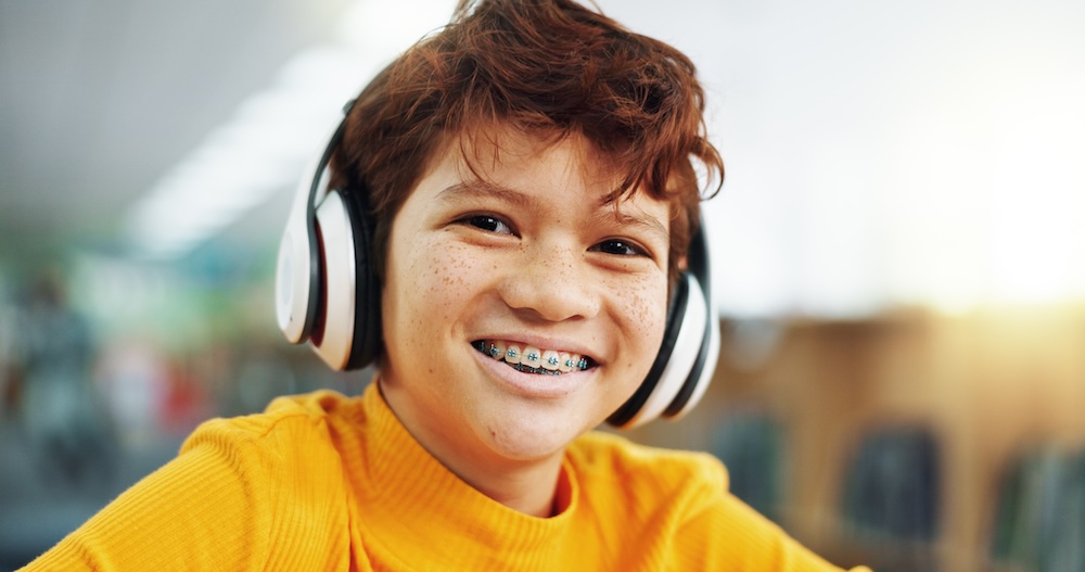 Smiling kid with braces wearing white headphones and a yellow shirt.