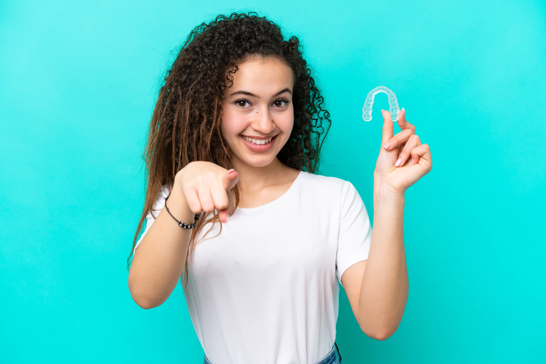 Smiling girl with curly hair, holding a clear retainer and pointing at the camera.
