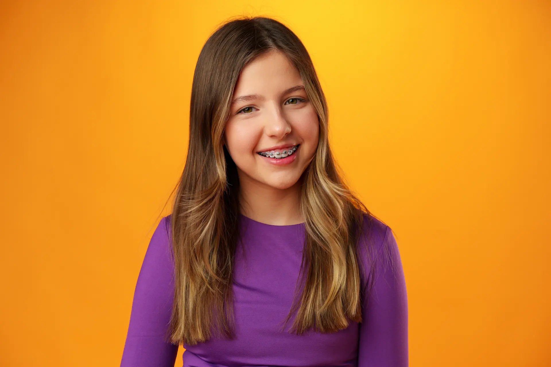 Smiling girl in a purple shirt wearing braces against an orange background.